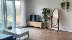 A modern living room with a large window, a white sofa, a coffee table, a TV on a wooden cabinet, a mirror, and a potted plant. - GLA Aewol (Jeju City)