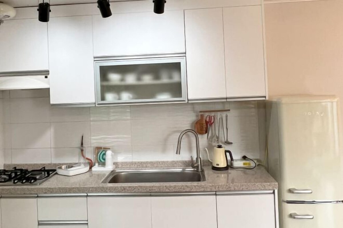 Una cocina con armarios blancos, un lavamanos de acero inoxidable, una estufa a gas y un refrigerador.