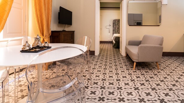 A hotel room with a dining area featuring a white table and clear chairs, a TV mounted on the wall, and a patterned floor. - Capo10 Liberty C1 in Syracuse (Syracuse)