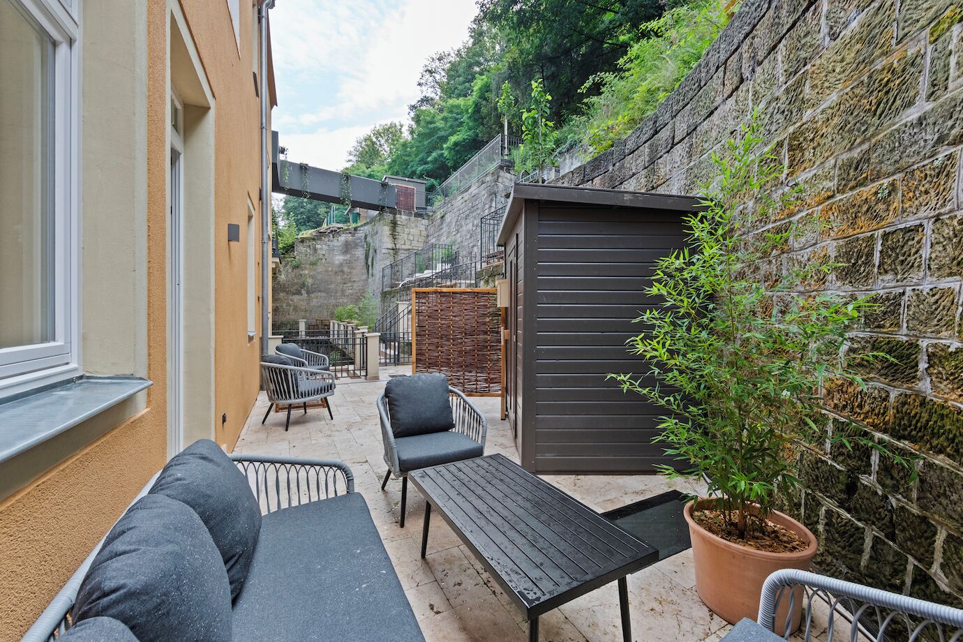 A narrow outdoor patio with a sofa, chairs, and a table. There is a small shed and a potted plant.