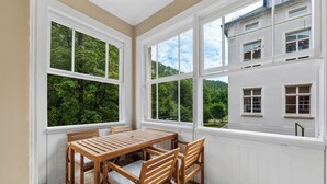 Quarto com mesa e cadeiras de madeira, janelas amplas com vista para o verde e paredes brancas.