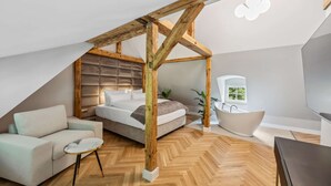 Un dormitorio con techo inclinado, estructura de techo de madera, cama, sofá, una pequeña mesa redonda, bañera y televisor.