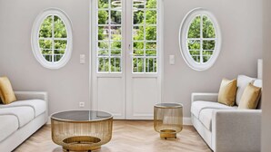 Una sala de estar moderna con dos sofás blancos, una mesa de centro de vidrio y una mesita auxiliar redonda dorada. Una puerta blanca con paneles de vidrio está centrada detrás de los sofás, y hay dos ventanas redondas a cada lado de la puerta.
