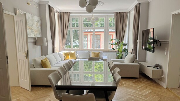 Ein modernes Wohnzimmer mit einer großen L-Form-Couch, einem Esstisch mit Stühlen und einem an der Wand befestigten Fernseher.