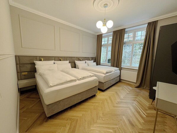 A hotel room with two beds, a wooden herringbone floor, a large window with curtains, and a ceiling light fixture.