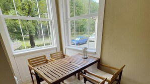 Ein Essbereich mit einem Holztisch und -stühlen, großen Fenstern mit Blick auf Bäume und parkende Autos sowie einem gefliesten Boden.