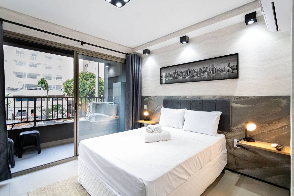 A modern hotel room with a large bed, a cityscape painting, a balcony with a view, and a small stool.