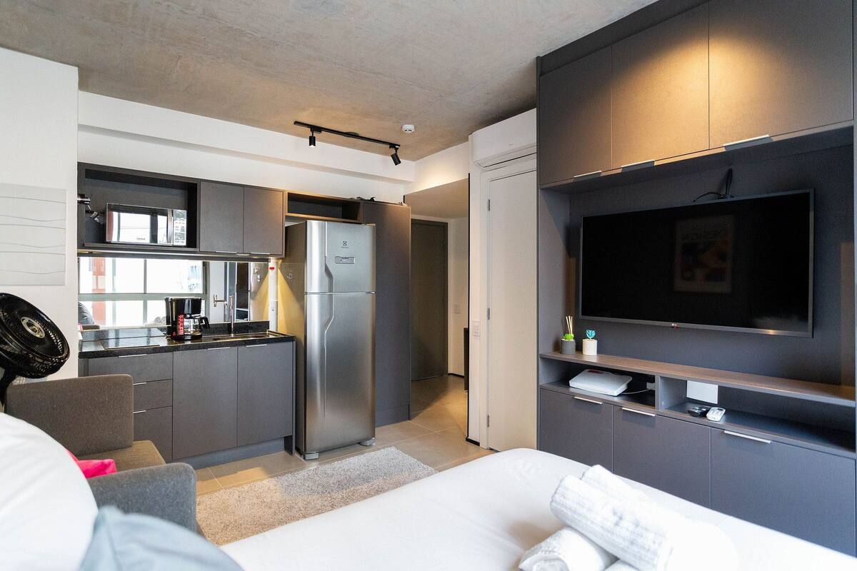 A modern hotel room with a kitchenette, a bed, a TV, and a seating area.