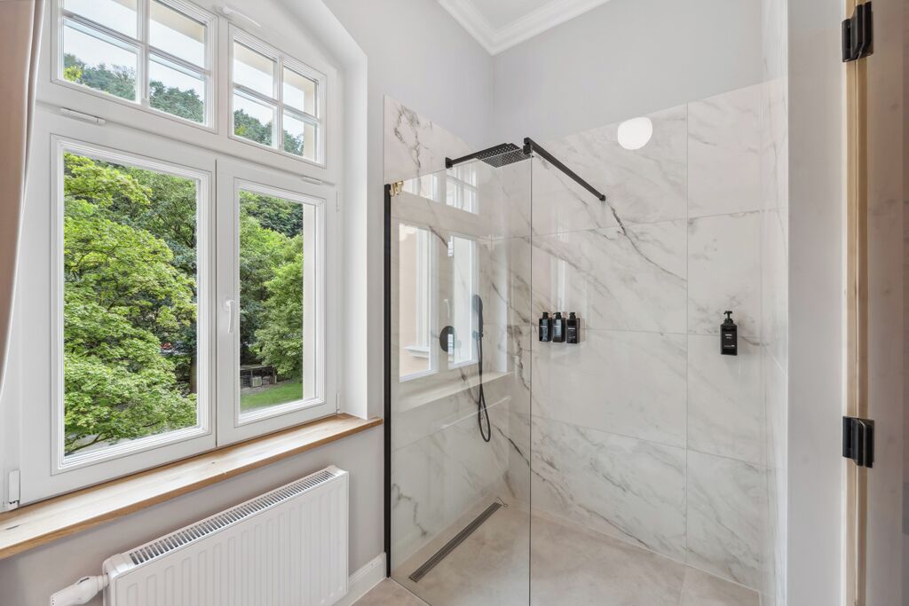 Une salle de bain moderne avec une douche à l’italienne en verre, une fenêtre donnant sur un espace verdoyant et un mur en marbre.