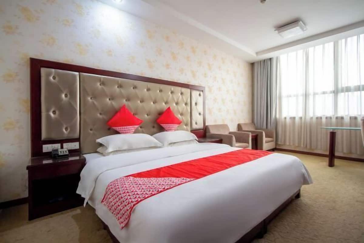 A hotel room with a large bed, a wooden headboard, two red pillows, a bedside table, a chair, and a window with curtains.