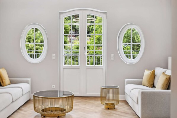 Uma sala de estar moderna com dois sofás brancos, uma mesa de centro de vidro e uma mesinha lateral redonda dourada. Uma porta branca com painéis de vidro está centralizada atrás dos sofás, e há duas janelas redondas de cada lado da porta.
