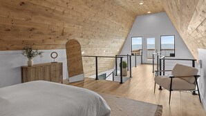 A bedroom with a sloped ceiling, wooden floor, a bed, a chair, and a view of the outdoors. - Mountain’s Edge by Appalachian A-Frames, Mountain View, Hot Tub, WiFi (Trenton)