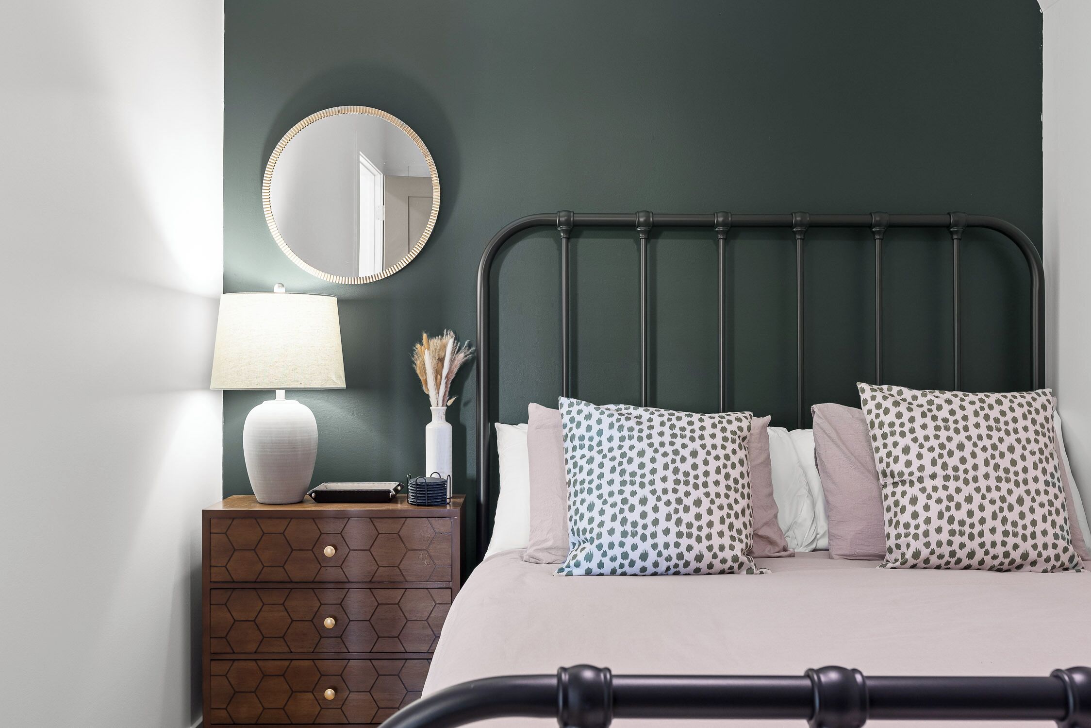 Une chambre à coucher bien rangée, avec un lit, une table de chevet dotée d’une lampe, un miroir et un vase décoratif.
