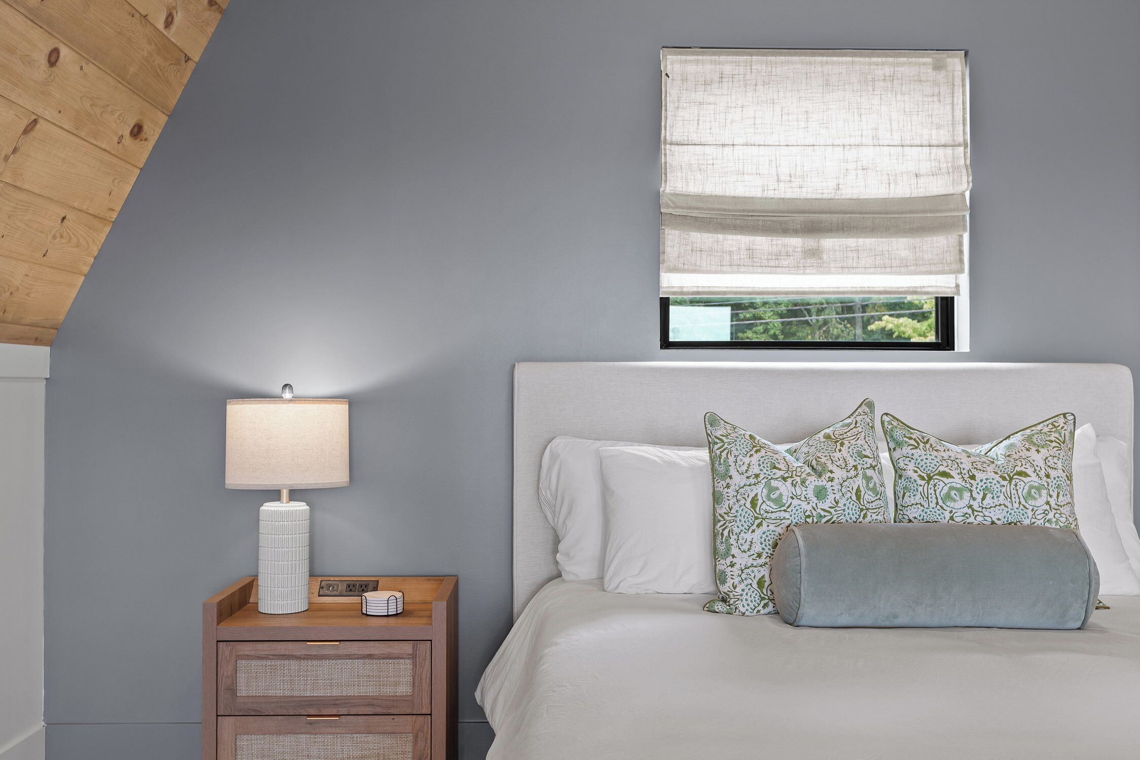 A bedroom with a bed, pillows, a nightstand with a lamp, and a window with a view.