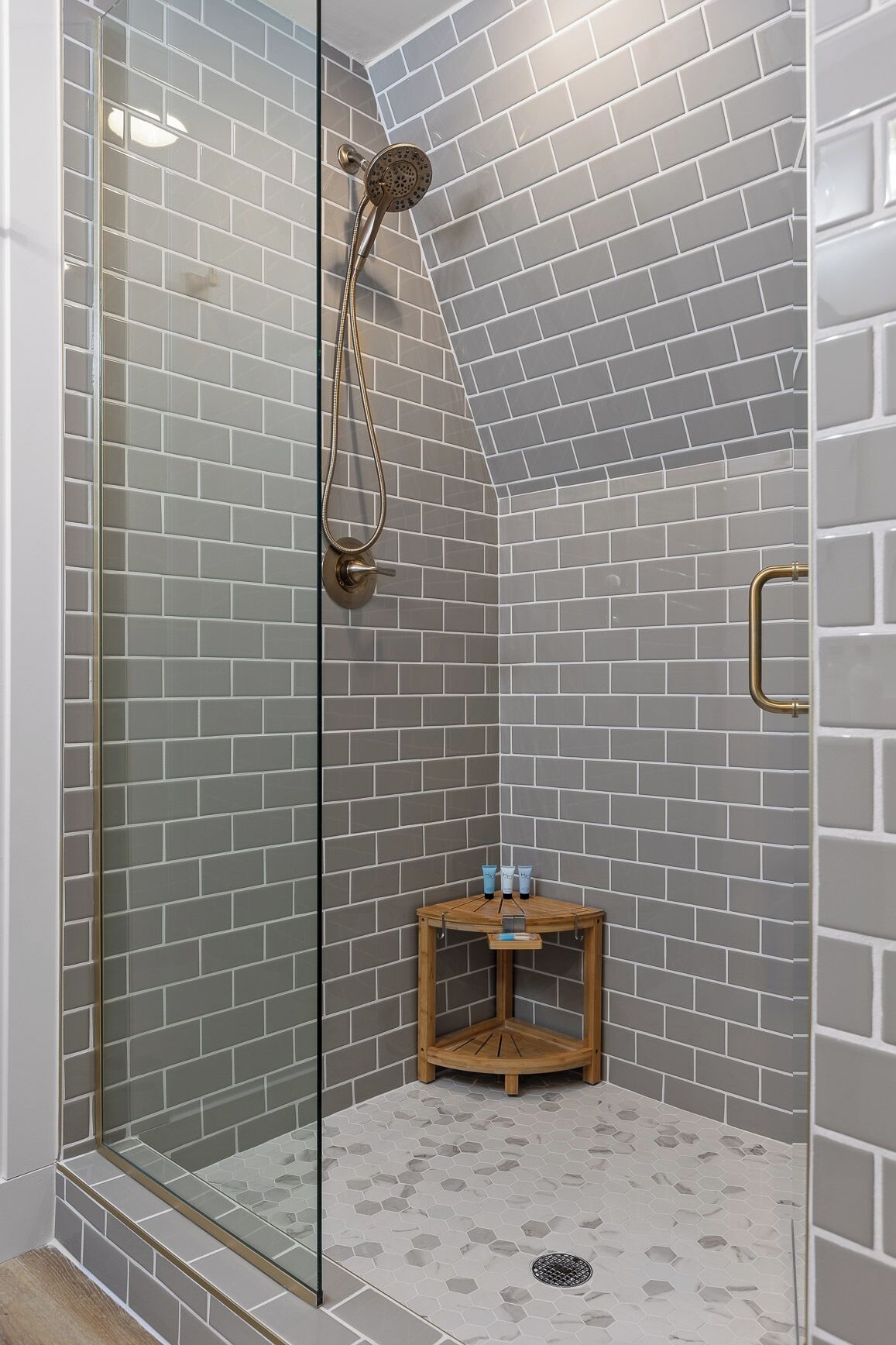 Eine Dusche mit Glastür, grauen Fliesen und einem kleinen Holzschemel mit Pflegeprodukten.