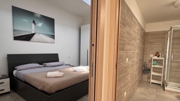 A modern bedroom with a bed, a skylight, a painting, and a bathroom with a shower and toilet. - Mungo in Fiumicino With 1 Bedrooms and 1 Bathrooms (Fiumicino)