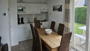 Une cuisine compacte avec des meubles blancs, un four à micro-ondes et une cuisinière à gaz. On y trouve une table à manger avec quatre chaises et un saladier posé dessus. Une fenêtre avec des rideaux est visible, et une petite table et un banc d’extérieur sont visibles à l’extérieur.