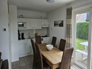 A compact kitchen with white cabinets, a microwave, and a gas stove. There is a dining table with four chairs and a bowl on it. A window with curtains is visible, and a small outdoor table and bench are seen outside.