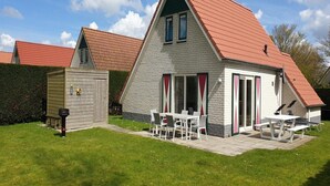 Une maison à deux étages avec un toit rouge, une terrasse dotée de mobilier d’extérieur et un petit abri de jardin.