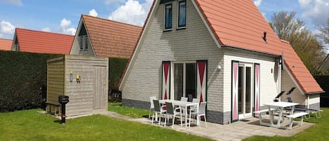 Een huis met twee verdiepingen, voorzien van een rood dak, een terras met tuinmeubilair en een kleine schuur.