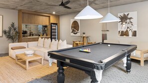 A pool table with balls, a sofa, a coffee table, a ceiling fan, pendant lights, and a billiards rack.