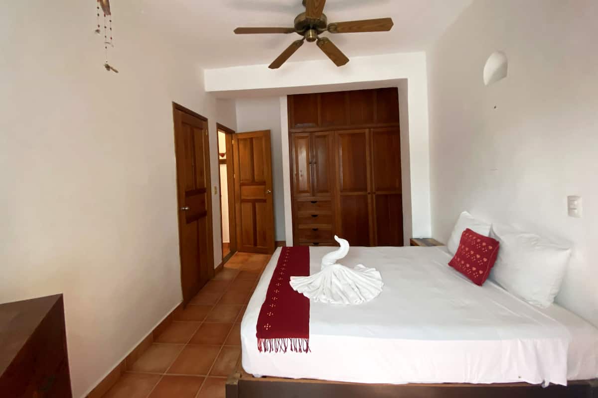 A bedroom with a bed, a ceiling fan, wooden doors, and a red pillow.