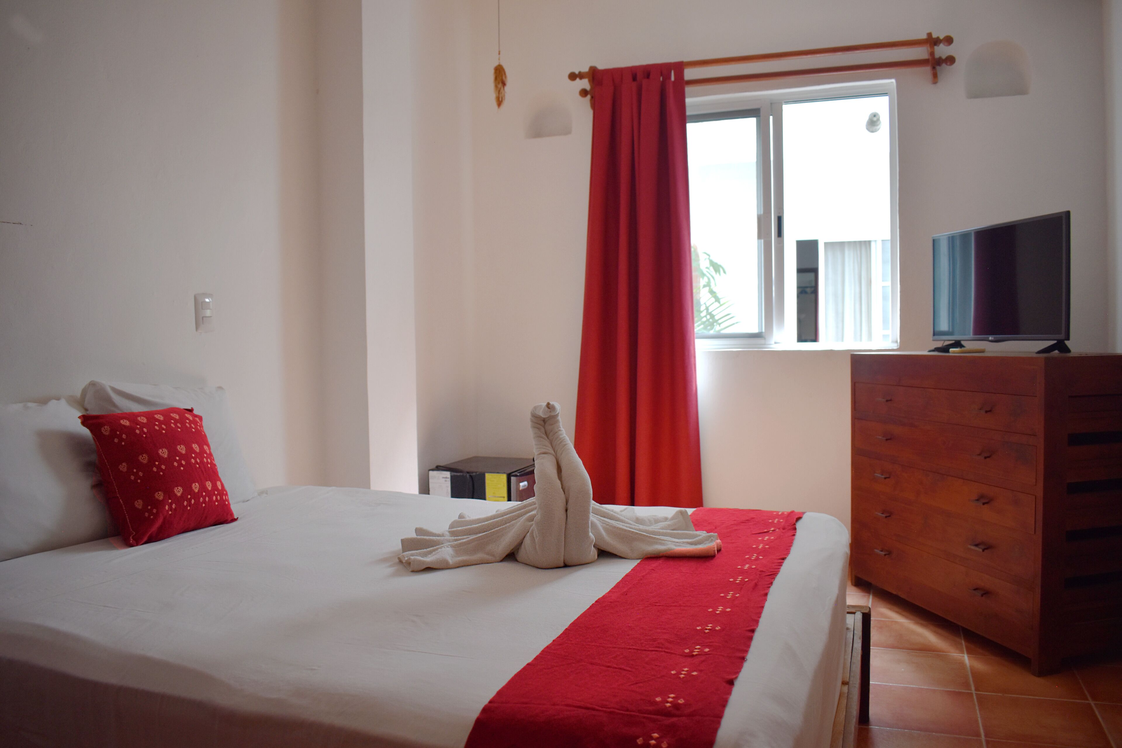 A neatly made bed with a red blanket and a towel arranged in a swan shape. There is a wooden dresser with drawers and a television on top.