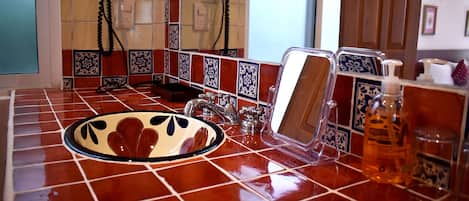 Un baño con piso de baldosas, un lavamanos con espejo, dispensador de jabón y un teléfono fijado a la pared.