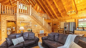 A wooden cabin interior with a staircase, two leather sofas, a dining table, and a kitchen area. - Smoky Traditions-Luxury Cabin,Gated,Pools,Mini-golf, Brand New Multi-Game Arcade (Sevierville)
