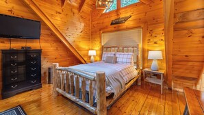 A cozy log cabin bedroom with a wooden bed, a TV, and a small table with a lamp. - Smoky Traditions-Luxury Cabin,Gated,Pools,Mini-golf, Brand New Multi-Game Arcade (Sevierville)