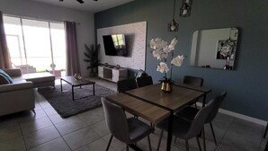 Una sala moderna con un sofá, una mesa de centro, una mesa de comedor y sillas. Un televisor de pantalla plana está montado en la pared, y hay una planta en maceta en el suelo.