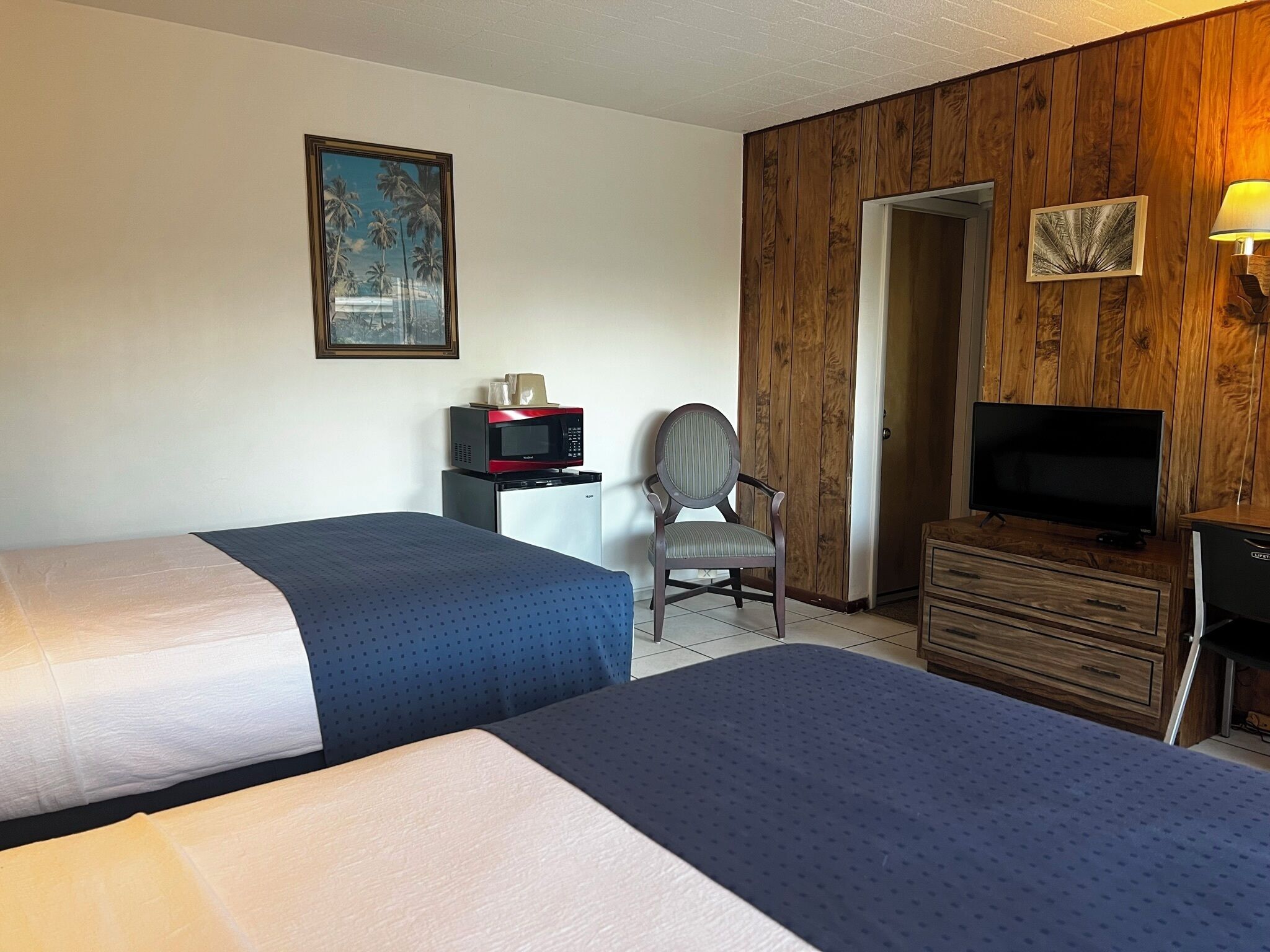 A hotel room with two beds, a desk with a microwave, a chair, a TV, and wooden paneling on the walls.