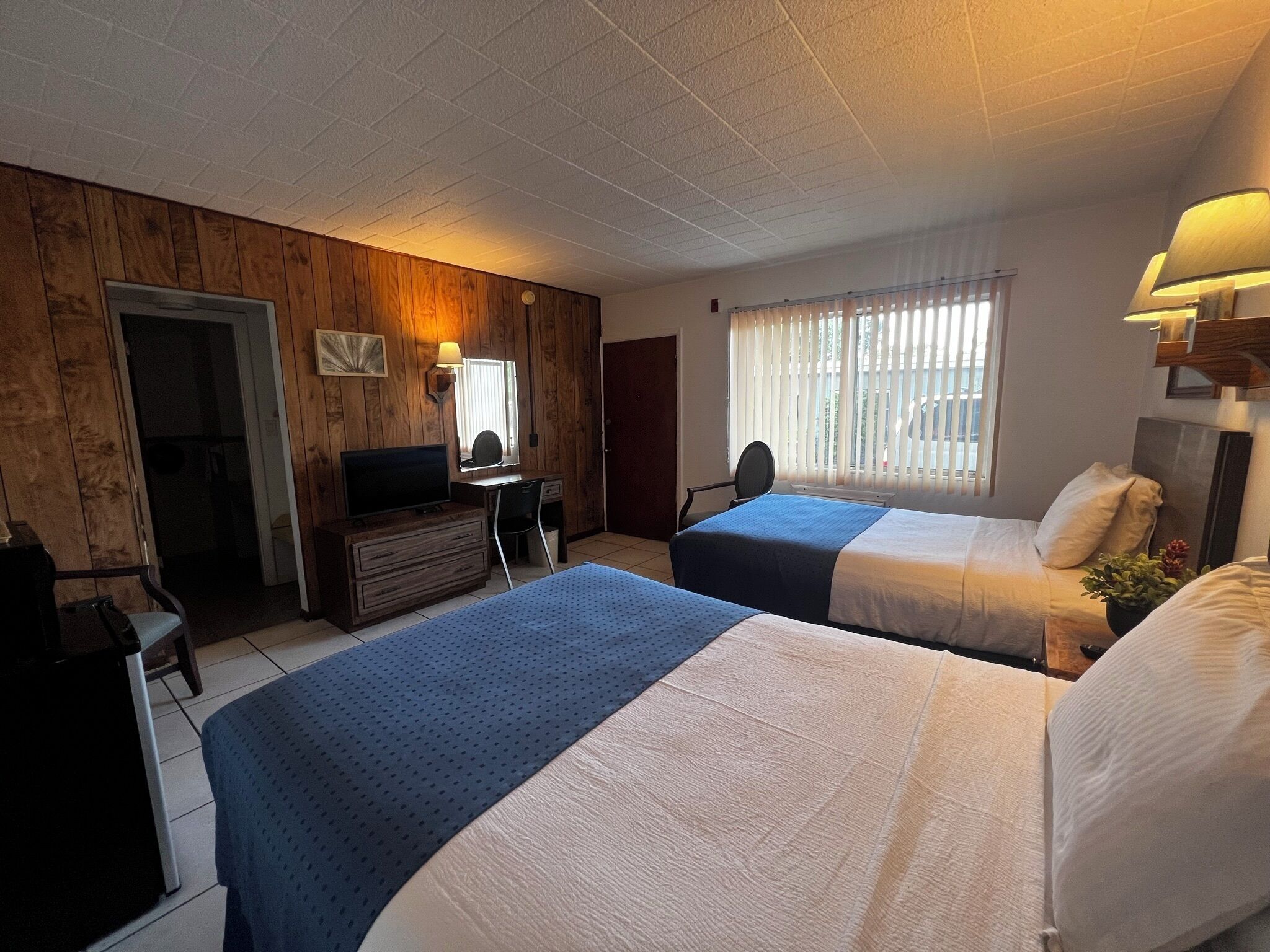 A hotel room with two beds, a desk with a computer, a television, and a window with blinds.