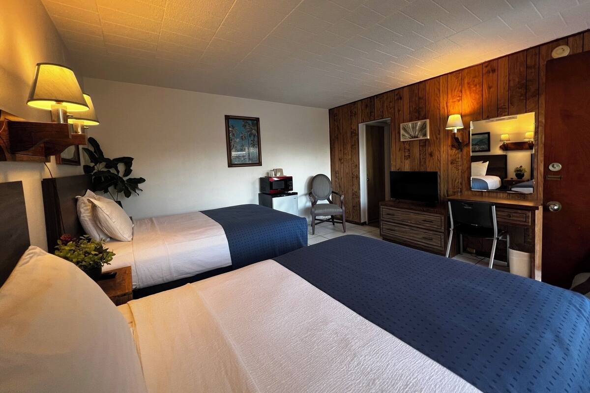 Ein Hotelzimmer mit zwei Betten, einem Schreibtisch mit Computer, einem Fernseher und einer Wand mit Holzpaneelen.