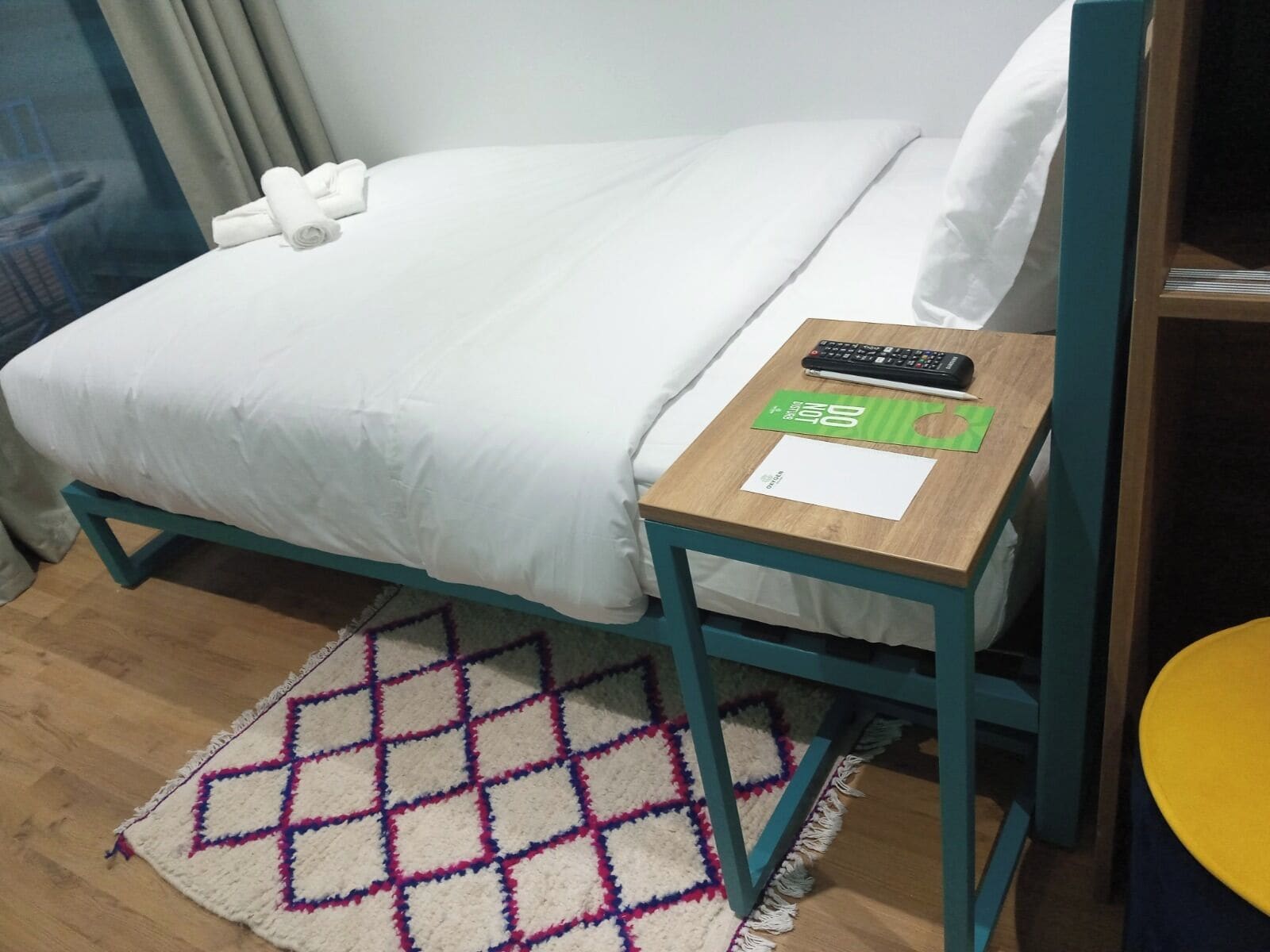 A neatly made bed with white linens, a wooden nightstand with a remote control and a green card, and a patterned rug on a wooden floor.