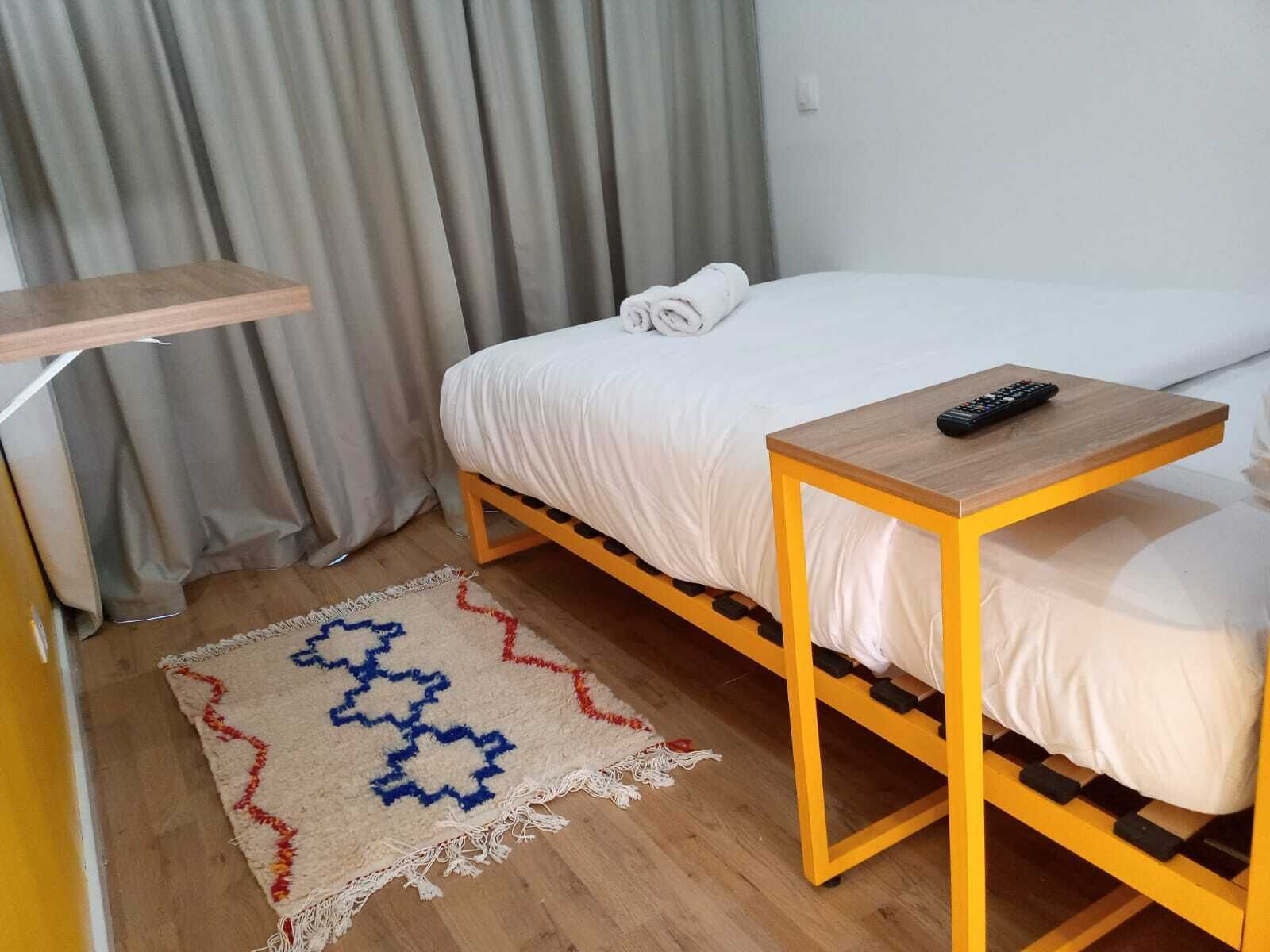 Uma cama de solteiro com estrutura de madeira, uma mesinha de cabeceira com controle remoto e um tapete estampado sobre piso de madeira.