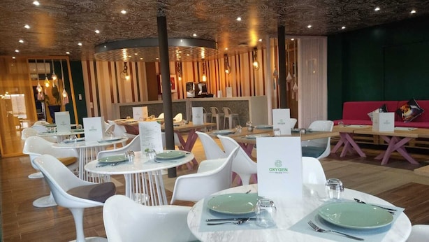 Moderni ravintola, jossa on pyöreät pöydät ja valkoiset tuolit. Ravintolassa on ainutlaatuinen kattorakenne ja taustalla vihreä seinä.