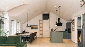A spacious living area with a high ceiling, a dining table, a green sofa, and a kitchen with a fireplace. - Luxury Pool House Near Sea - By Traum Ferienwohnungen (Ulfborg)