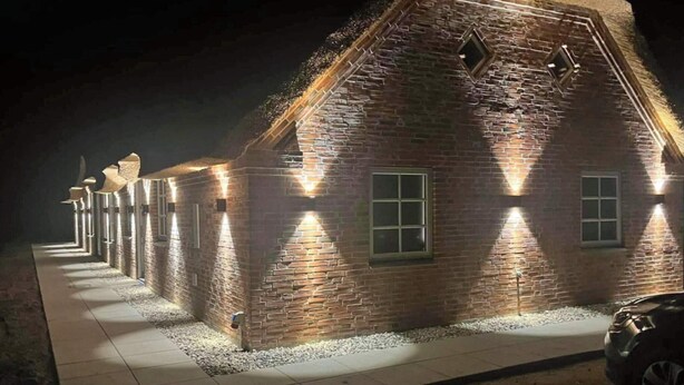 A brick building with a thatched roof, illuminated by wall-mounted lights, and a paved walkway leading to it. - Luxury Pool House Near Sea - By Traum Ferienwohnungen (Ulfborg)