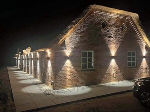 Un bâtiment en briques surmonté d’un toit de chaume, éclairé par des lampes fixées aux murs, et un chemin pavé menant à l’entrée.