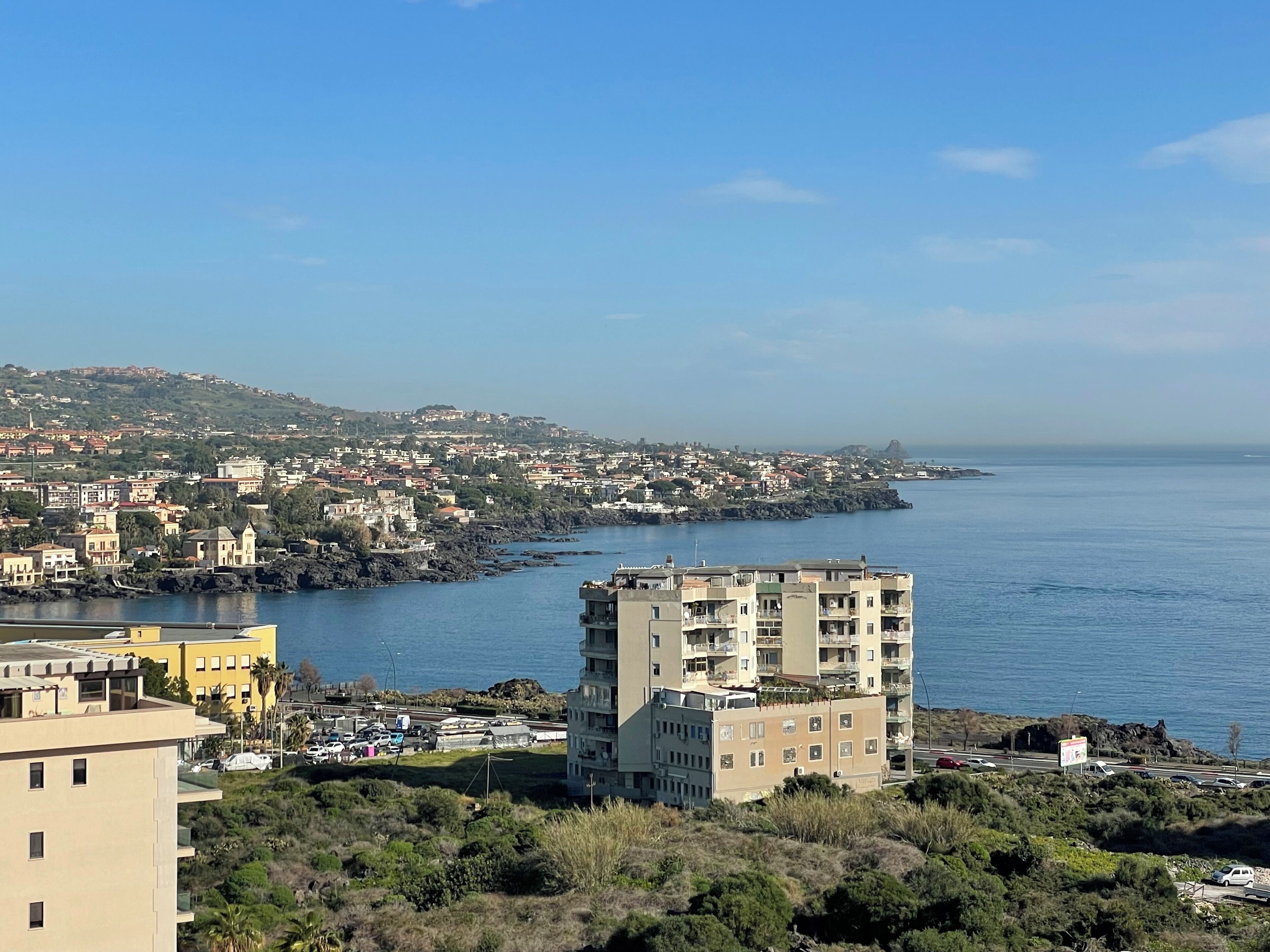 Un paysage urbain côtier avec, au premier plan, un immeuble imposant à plusieurs étages, un port de plaisance avec des bateaux, et, à l’arrière-plan, une colline densément bâtie.