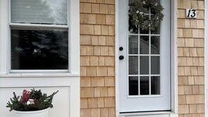Une maison avec une porte blanche, un revêtement extérieur en bardeaux de bois, une grande fenêtre et un pot de fleurs contenant une plante aux baies rouges.