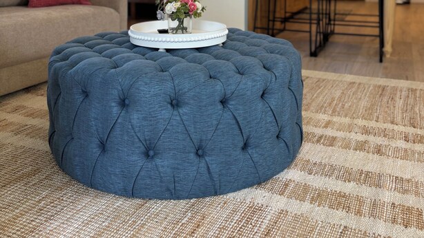 Un salon comprenant un pouf capitonné, un canapé et une table basse agrémentée d’un arrangement floral.