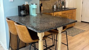 Une cuisine avec des meubles en bois, un plan de travail en granit, deux tabourets de bar, une machine à café et un évier.