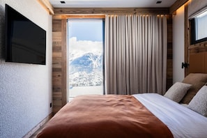 A bedroom with a large bed, a TV mounted on the wall, and a view of mountains through a window.