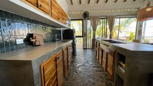 A kitchen with wooden cabinets, a stone floor, and a view of palm trees through large windows. - Charming oceanfront cabin with stunning views, private terrace, and beach access (Santa Marta)