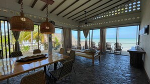 A spacious dining area with a view of the beach, featuring a wooden table, wicker chairs, and a hanging light fixture. - Charming oceanfront cabin with stunning views, private terrace, and beach access (Santa Marta)