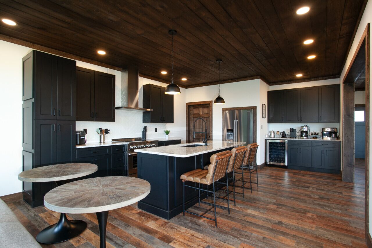 A modern kitchen with dark cabinetry, a central island with bar stools, and a dining area with round tables.