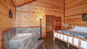 Une chambre aménagée dans un chalet en rondins, avec un plafond en bois, un grand lit, un banc en bois, une baignoire et une porte donnant sur une autre pièce.