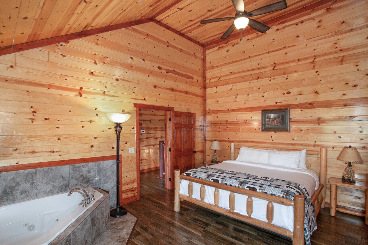 A log cabin bedroom with a wooden ceiling, a single bed, a nightstand, a lamp, a door, and a bathtub.